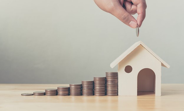 Mão coloca moeda sobre pilhas crescentes ao lado de casa em miniatura, ilustrando poupança e gestão financeira.