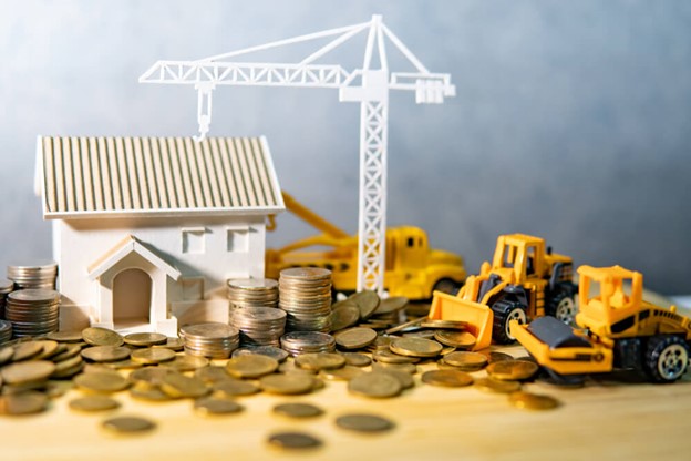 Casa em miniatura, guindaste de brinquedo e pilhas de moedas representam construção e crescimento de patrimônio.