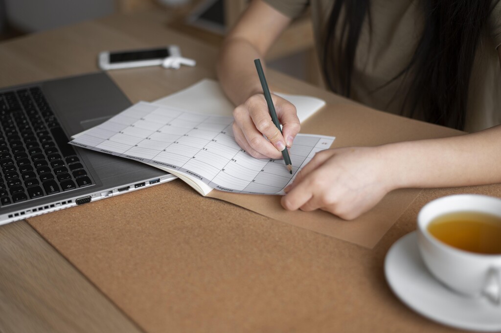 Pessoa preenchendo cronograma de estudos em calendário mensal ao lado de notebook, organizando a rotina de aprendizagem.