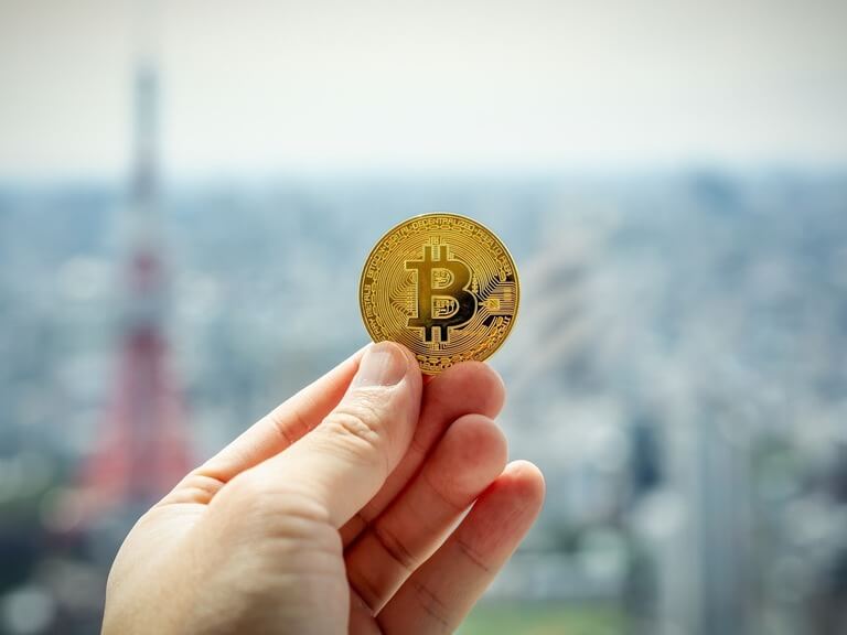 Mão segurando uma moeda de bitcoin e atrás existe uma vista de cidade