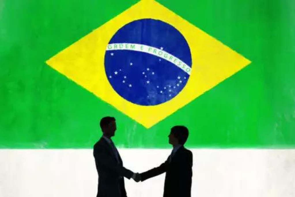 Políticas públicas brasileiras sendo representadas por um aperto de mãos. Atrás, a bandeira do Brasil