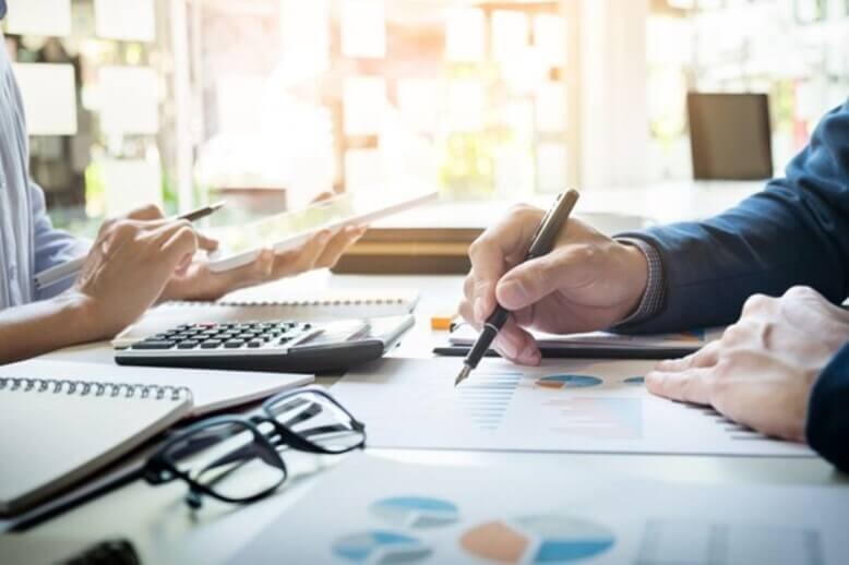 Pessoas analisam gráficos e documentos em uma mesa, com calculadora, cadernos e óculos ao lado.