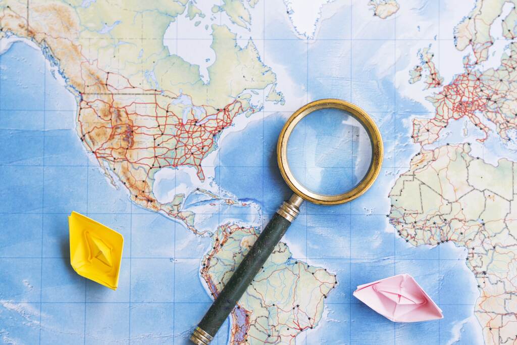 Lupa sobre mapa-múndi e barcos de papel representam a análise das relações internacionais e do poder global.