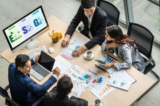 Equipe reunida em mesa de reunião discutindo relatórios e gráficos, com monitor exibindo ícones de ESG ao fundo