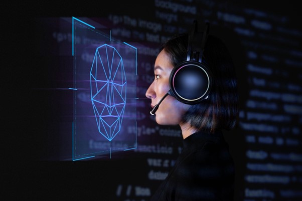 Mulher usando fones de ouvido com microfone, olhando para um holograma de rosto digital composto por linhas e pontos, exibido em um fundo escuro com códigos de programação ao fundo. A imagem ilustra a interação com a tecnologia de deepfake.