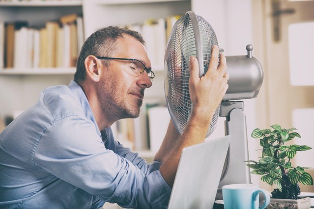 Homem sentado em uma mesa de escritório, inclinado próximo a um ventilador, tentando se refrescar em um ambiente quente e fugir da ebolição global