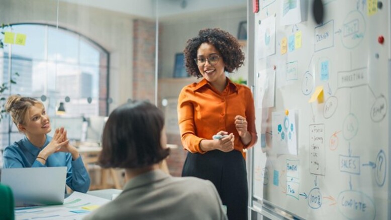 Uma mulher discute projetos com colegas diante de um quadro branco, evidenciando os valores de uma empresa