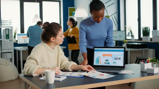 Duas profissionais discutindo estratégias de marketing em frente a um laptop aberto, em um ambiente de escritório movimentado