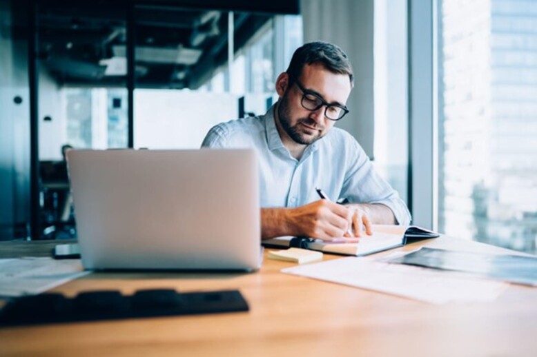 Homem com especializações em Administração escreve em um caderno, concentrado em sua mesa de trabalho com laptop e anotações.