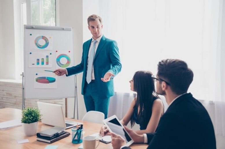 Homem com especializações em Administração conduz uma apresentação em um ambiente corporativo, explicando gráficos para colegas atentos. Eles discutem estratégias com tablets e laptops sobre a mesa.