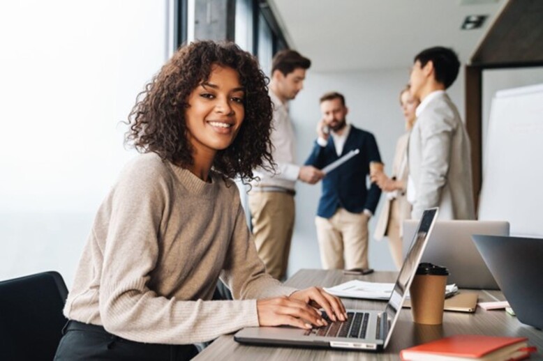 Mulher sorridente com especializações em Administração trabalha em um laptop em um escritório moderno. Ao fundo, colegas discutem estratégias em uma reunião informal.