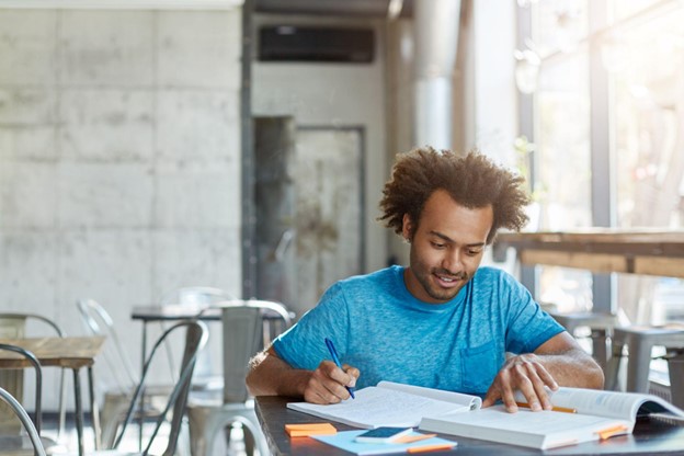 Estudante estudando em uma biblioteca, com livros na mesa, simbolizando a jornada de quatro anos da graduação em Economia