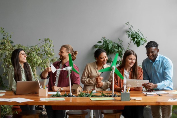 Grupo diverso de profissionais reunidos em torno de uma mesa, discutindo ideias sobre sustentabilidade empresarial, com miniaturas de turbinas eólicas e plantas ao fundo, simbolizando práticas ecológicas e inovação.