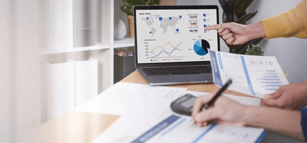 Duas pessoas analisam gráficos e relatórios de contabilidade financeira em um laptop e documentos impressos, destacando dados de desempenho e tendências de mercado.