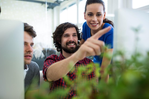 Três profissionais colaborando em frente a um computador, com plantas ao redor, simbolizando sustentabilidade empresarial e inovação em projetos corporativos.