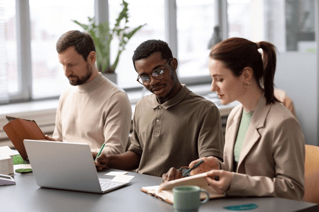 Três pessoas em um escritório colaboram em um projeto, utilizando um laptop e cadernos, demonstrando habilidades profissionais em um ambiente de trabalho moderno.