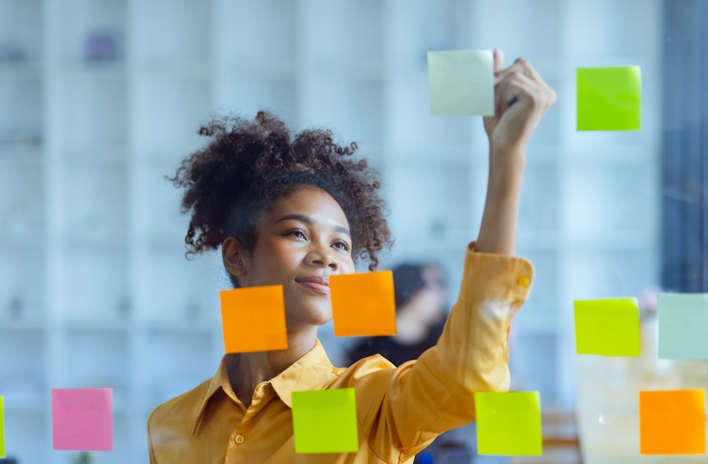 Profissional organiza post-its em quadro de planejamento, demonstrando proatividade, autonomia e iniciativa no trabalho.